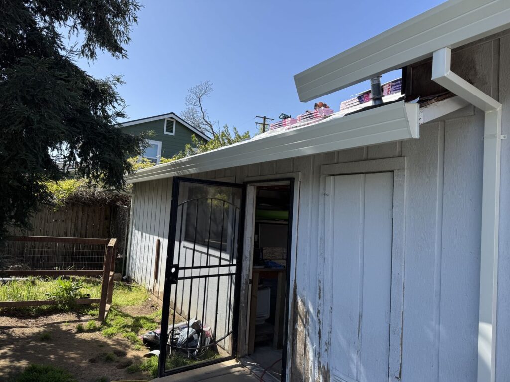 Back of a white home with newly installed white rain gutters and downspouts under a clear blue sky. The home has a black screen door that is open, a closed wooden door, and some construction materials on the roof. There is a fenced area with grass and dirt beside the home, and a large evergreen tree