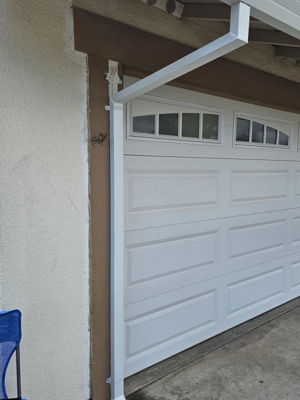 White downspout installed on the left side of a white garage door. The white downspout is attached to white gutters, which was also installed.
