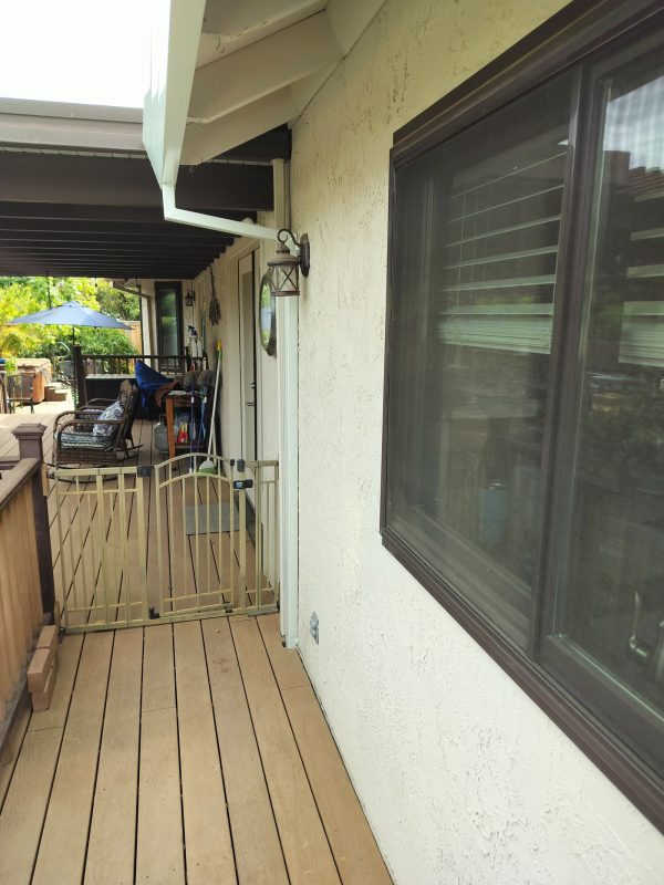 A white downspout following down the side of wall. The side of the deck extends with a patio cover, covering a part of the deck.