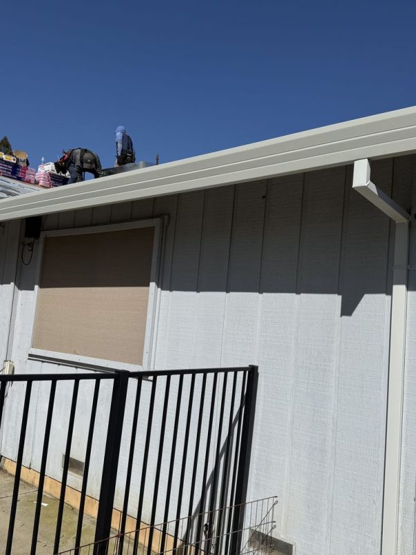 Side of a white home with newly installed white rain gutters and downspout under a clear blue sky. The home has a black fence. Some construction materials on the roof.