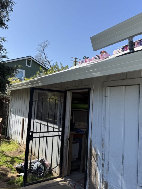 Back of a white home with newly installed white rain gutters and downspouts under a clear blue sky. The home has a black screen door that is open, a closed wooden door, and some construction materials on the roof. There is a fenced area with grass and dirt beside the home, and a large evergreen tree