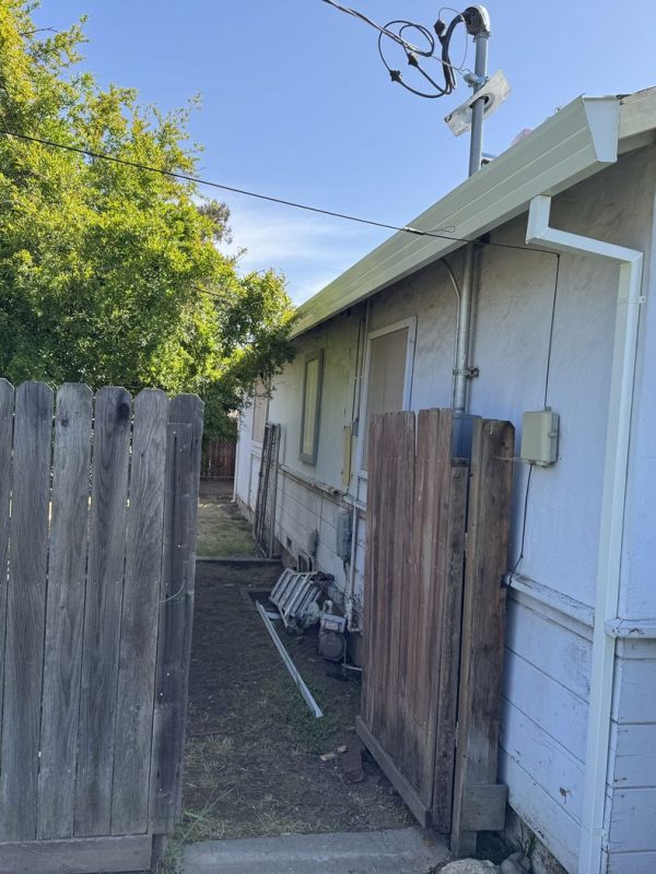 Side of a white home with newly installed white rain gutters and downspouts under a clear blue sky. A brown fence is beside the home, featuring the fence door open. Beside the home, and a large evergreen tree
